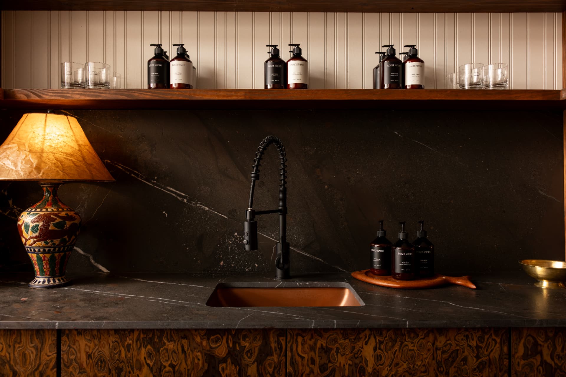 A washstand inside a Ranger Station house — hand wash and hand cream bottles lined along a stone counter beneath a wooden shelf of rocks glasses.