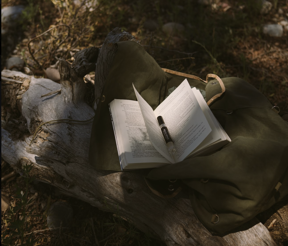 An open book and a Ranger Station perfume vial resting on a fallen log beside a canvas rucksack in the woods.
